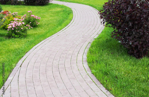 Curved paved with bricks footpath running ahead along a green grassy lawn. Landscaping design