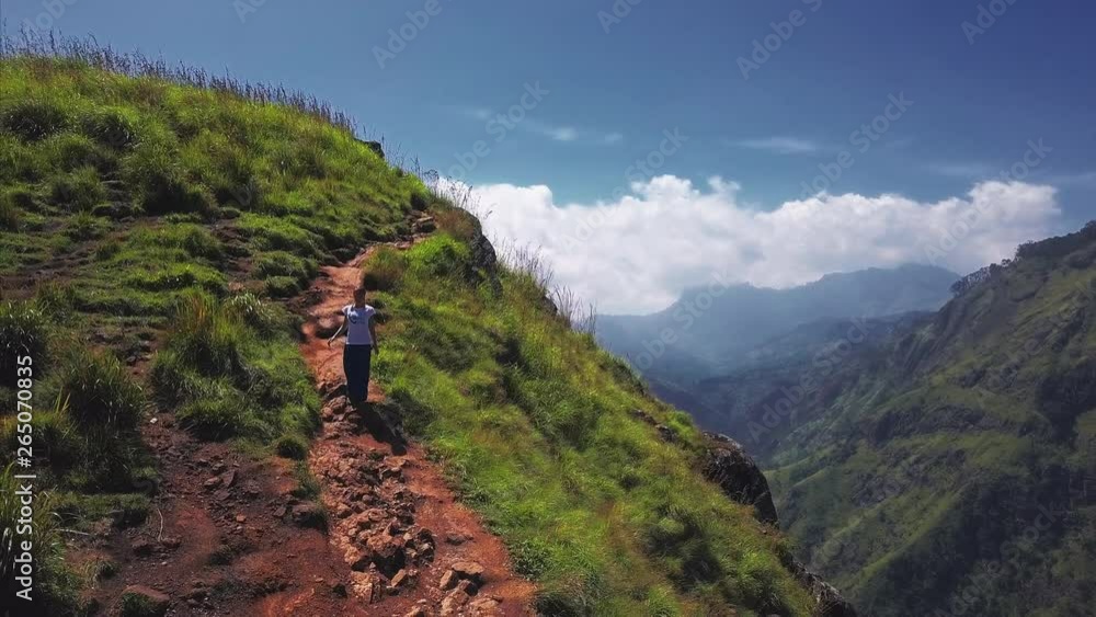 Woman walks on the mountain trail lying on the edge of the cliff with stunning valley views. The place named Little Adam's Peak in Ella, Sri Lanka Woman walks on the mountain trail lying on the edge of the cliff with stunning valley views. The place named Little Adam's Peak in Ella, Sri Lanka