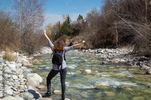 Young beautiful girl on nature with a backpack on the river bank. Good mood on a bright spring day. Against the background of the forest and a beautiful mountain river. Tourism and travel.