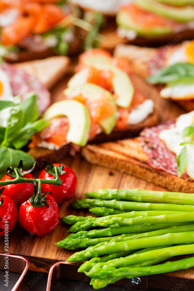 Huge healthy breakfast with sun-dried tomatoes, sandwiches with scrambled eggs, sausage, salmon, arugula, curd cheese, avocado on a wooden board