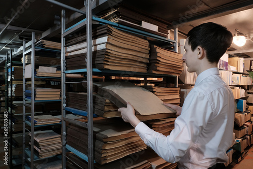 A student in the library or in the archive room is looking for information. A schoolboy in a white shirt pulls out an old book from a bookshelf. Concept back to school or study of historical documents