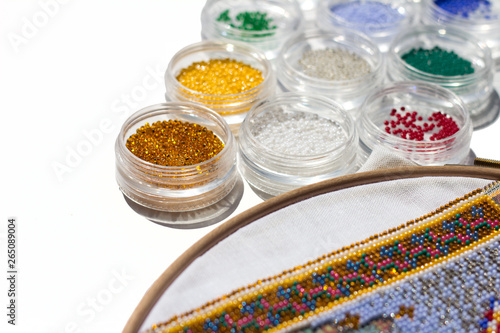 Embroidery. Round transparent containers with a bead next to a fixed canvas on the embroidery frame on which there is a print with symbols and a part of the ornament already embroidered with beads.