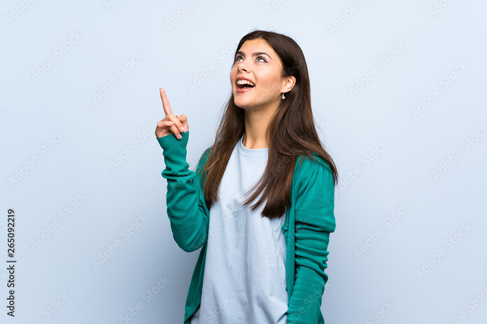 Teenager girl over blue wall intending to realizes the solution while lifting a finger up Teenager girl over blue wall intending to realizes the solution while lifting a finger up