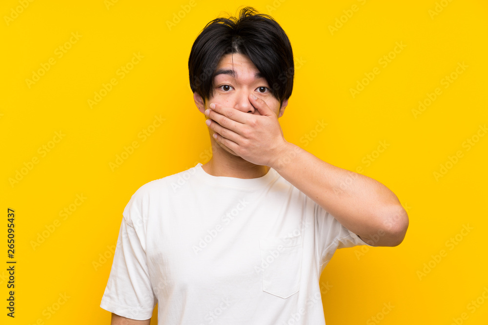 Asian man over isolated yellow wall covering mouth with hands Asian man over isolated yellow wall covering mouth with hands
