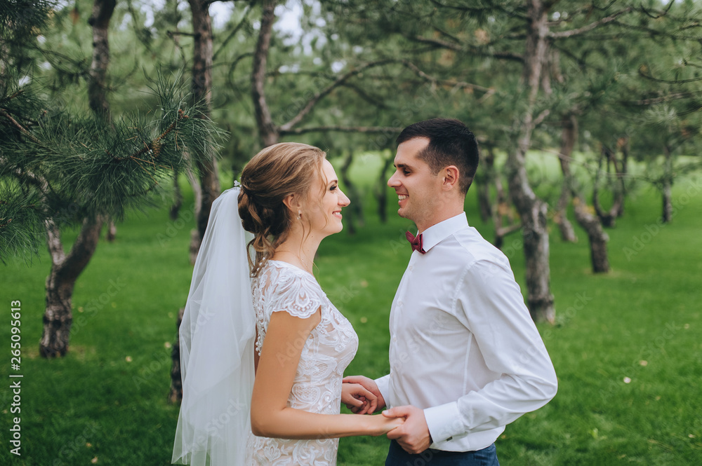 Beautiful newlyweds tenderly embrace in the forest against the background of green grass and pines. Wedding portrait of a stylish bride and a cute curly bride in a white dress.