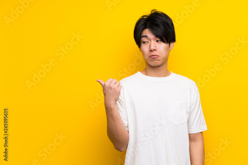 Asian man over isolated yellow wall unhappy and pointing to the side