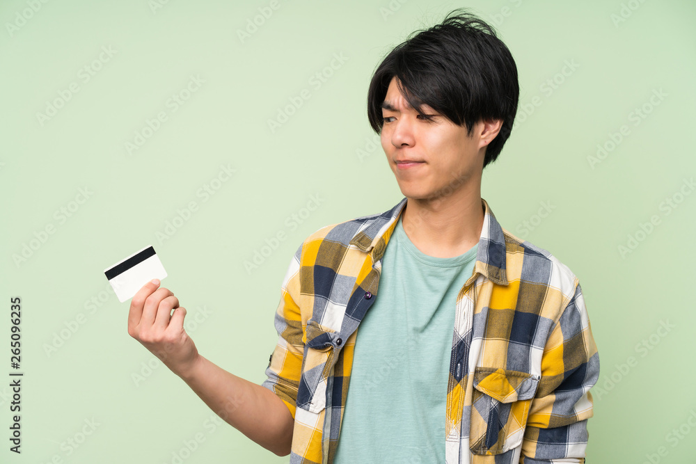 Asian man over isolated green wall holding a credit card Asian man over isolated green wall holding a credit card