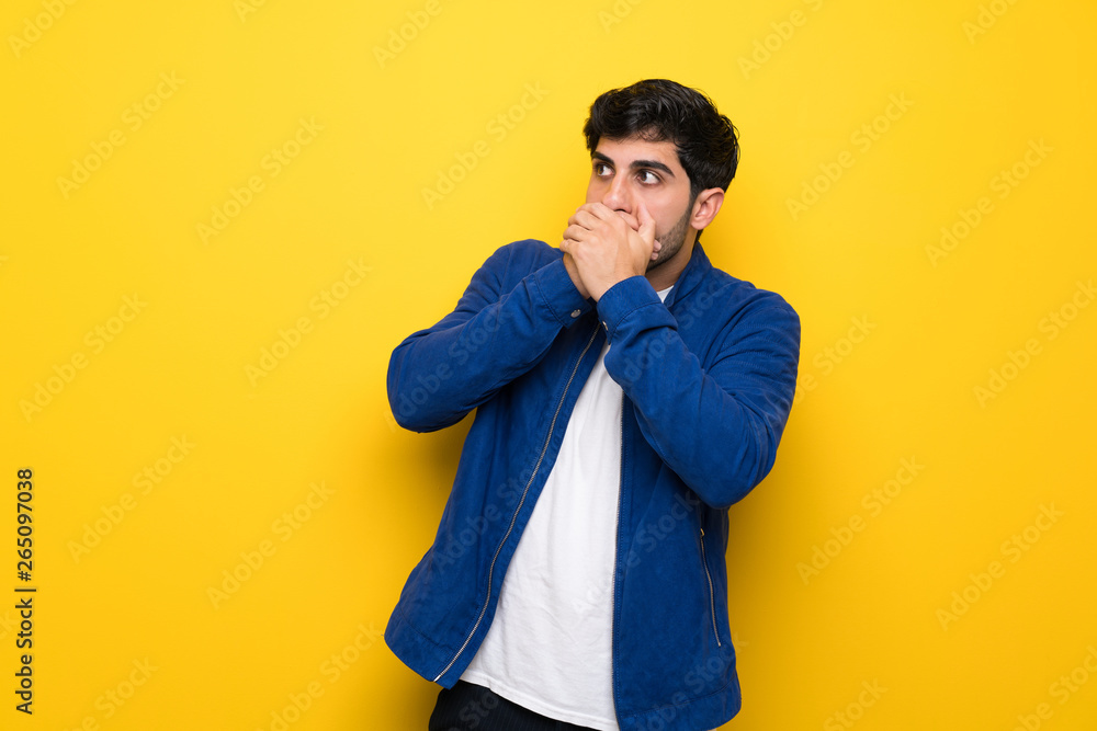 Man with blue jacket over yellow wall covering mouth and looking to the side Man with blue jacket over yellow wall covering mouth and looking to the side