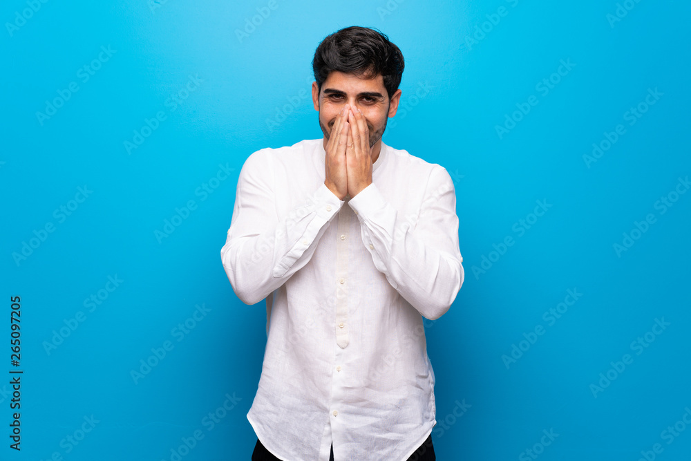 Young man over isolated blue wall smiling a lot while covering mouth Young man over isolated blue wall smiling a lot while covering mouth