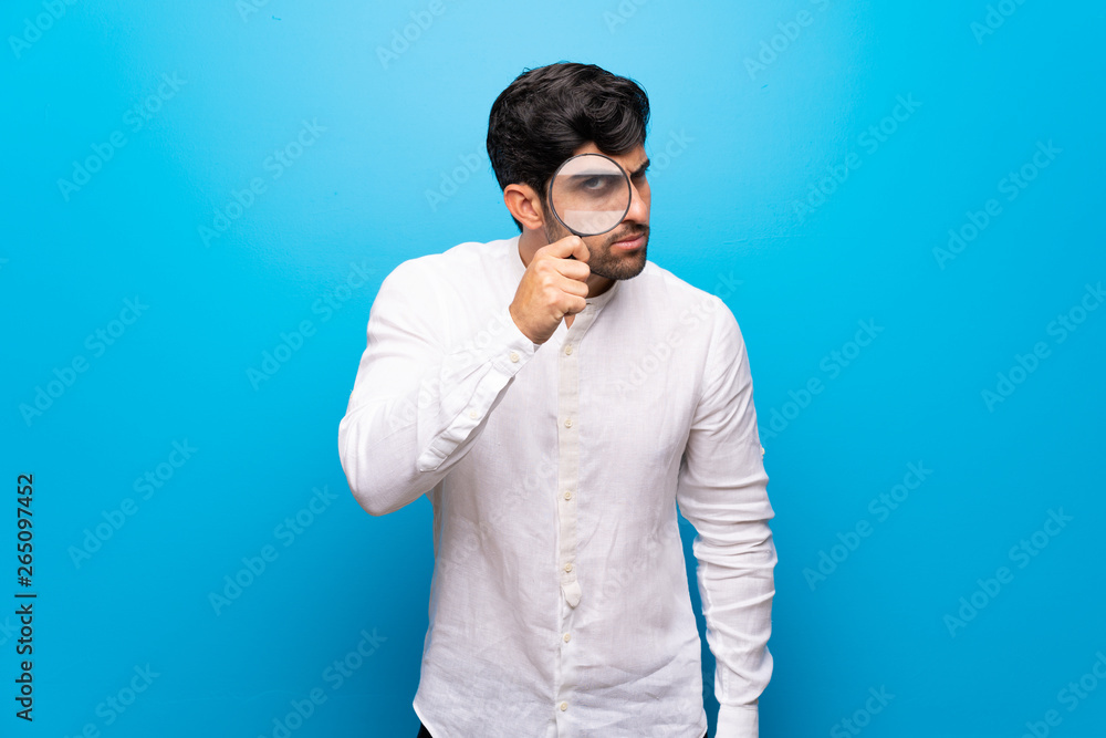 Young man over isolated blue wall taking a magnifying glass and looking through it