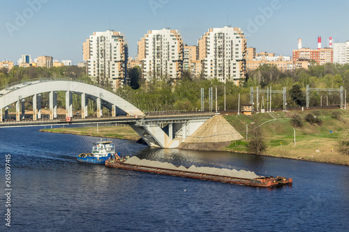 Tugboat pushes a barge laden with gray gravel. Against the background of the railway bridge over the canal, forests and urban development.