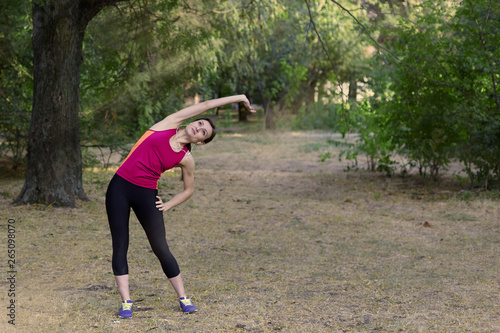 Fototapeta Naklejka Na Ścianę i Meble -  Gorgeous young slim woman makes gym in morning park. Side tilt, bright sportswear, white earphones, calmness and relax atmosphere, stretching training. Outdoors, copy space.