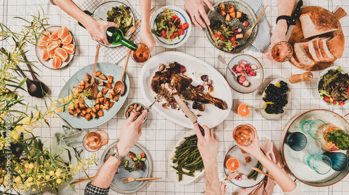 Family or friends gathering dinner. Flat-lay of hands of people eating roasted lamb shoulder, salads, vegetables, drinking rose wine over white checkered tablecloth, top view. Celebration party dinner