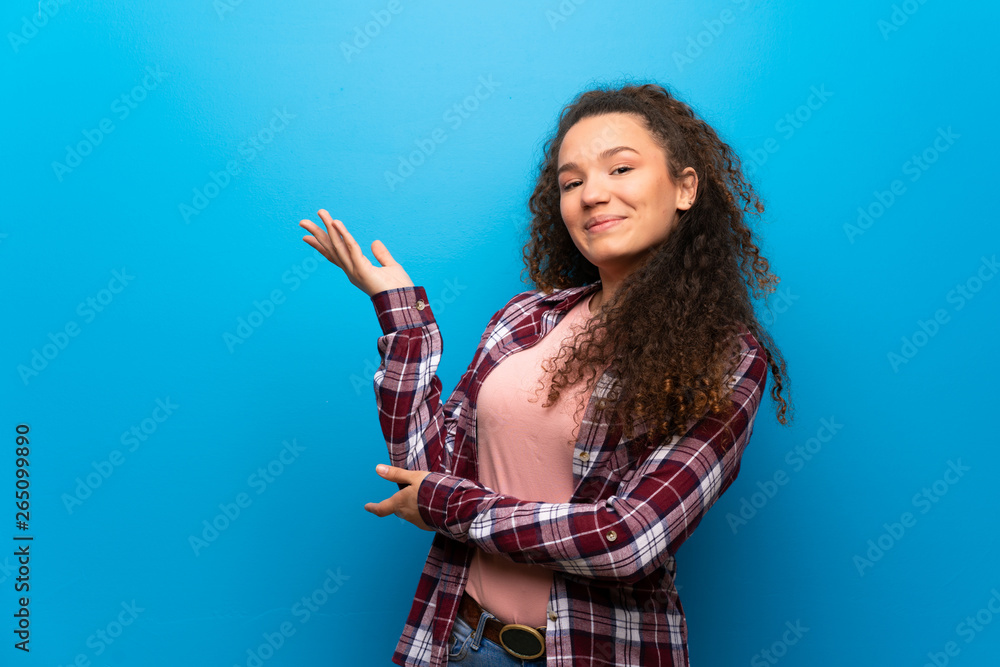 Teenager girl over blue wall extending hands to the side for inviting to come Teenager girl over blue wall extending hands to the side for inviting to come