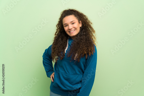 Teenager girl over green wall posing with arms at hip and smiling