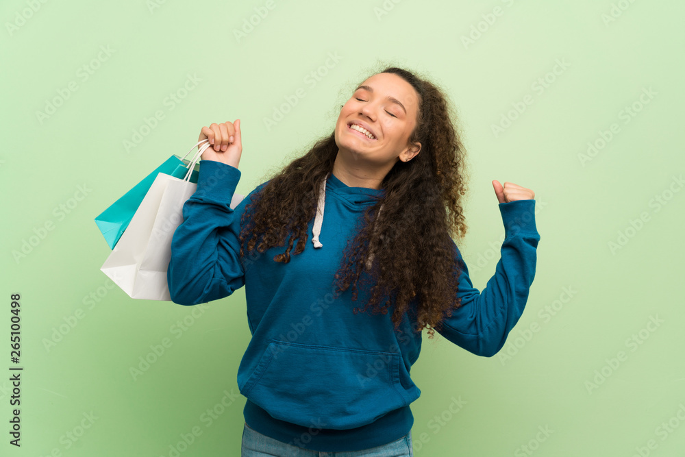 Teenager girl over green wall holding a lot of shopping bags in victory position Teenager girl over green wall holding a lot of shopping bags in victory position