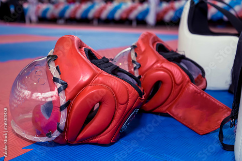 Two red karate fighting helmet with clear plastic masks and two body protective vests on the floor.