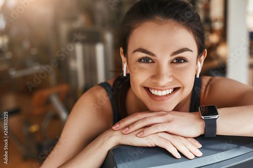 Fotografi Always in good mood! Portrait of cute and cheerful woman with smart watch on her hand looking at camera with toothy smile while taking a rest at gym