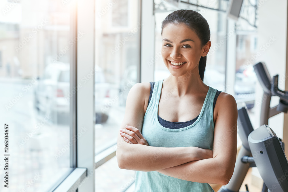 Sport is my lifestyle. Young and cheerful woman in sportswear looking at camera with smile while standing in front of windows at gym