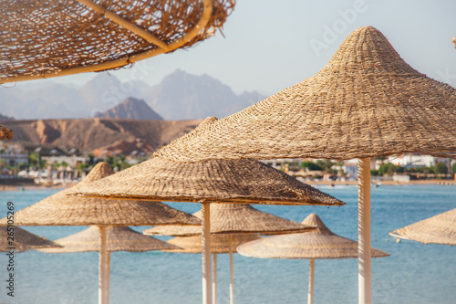 A row of straw umbrellas to protect against overheating on a sandy beach against a blue sky and blue sea. The concept of summer holidays, tourism, travel.