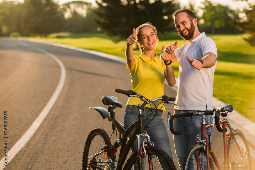 Joyful couple of cyclists gesturing thumbs up. Happy smiling people giving thumbs up sign outdoors. Summer active rest. Joyful couple of cyclists gesturing thumbs up. Happy smiling people giving thumbs up sign outdoors. Summer active rest.