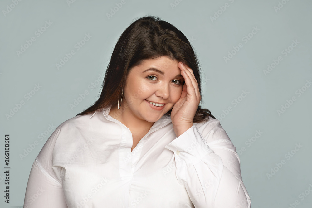People, beauty and lifestyle concept. Isolated shot of beautiful charming young brunette plus size woman wearing round earrings and formal shirt having shy timid look, hiding face behind her hand People, beauty and lifestyle concept. Isolated shot of beautiful charming young brunette plus size woman wearing round earrings and formal shirt having shy timid look, hiding face behind her hand