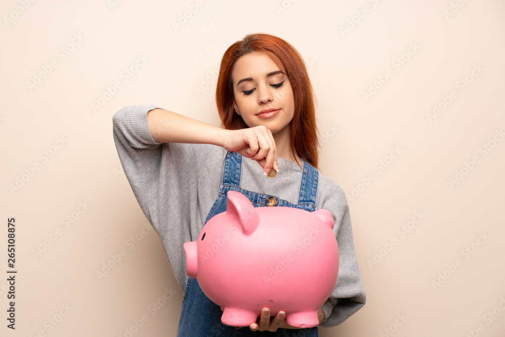 Young redhead woman over isolated background taking a piggy bank and happy because it is full Young redhead woman over isolated background taking a piggy bank and happy because it is full
