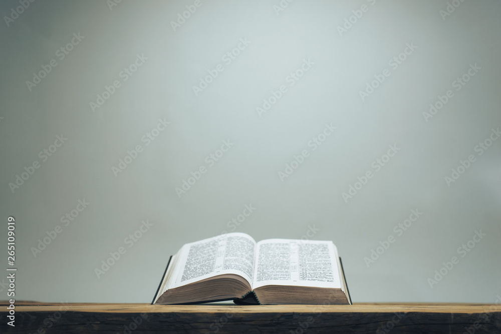 Open Holy Bible on a old oak wooden table.  Grey wall background.. - obrazy, fototapety, plakaty 