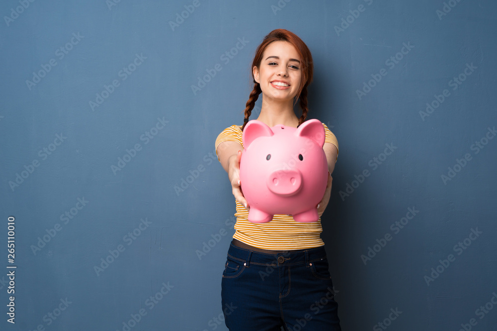 Young redhead woman over blue background holding a piggybank - obrazy, fototapety, plakaty 