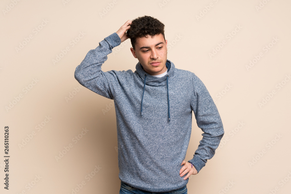 Young man over isolated wall having doubts while scratching head Young man over isolated wall having doubts while scratching head