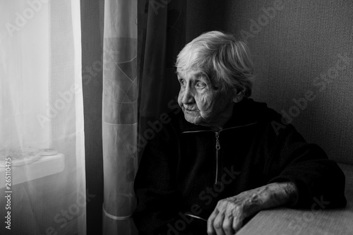 An elderly woman in his house looks out the window longingly. Old lady retired. Black and white photo.