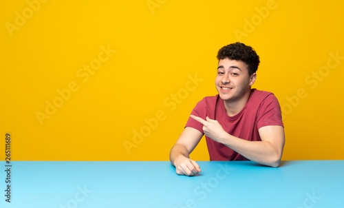 Young man with colorful wall and table pointing to the side to present a product
