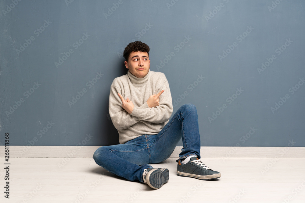 Young man sitting on the floor pointing to the laterals having doubts Young man sitting on the floor pointing to the laterals having doubts