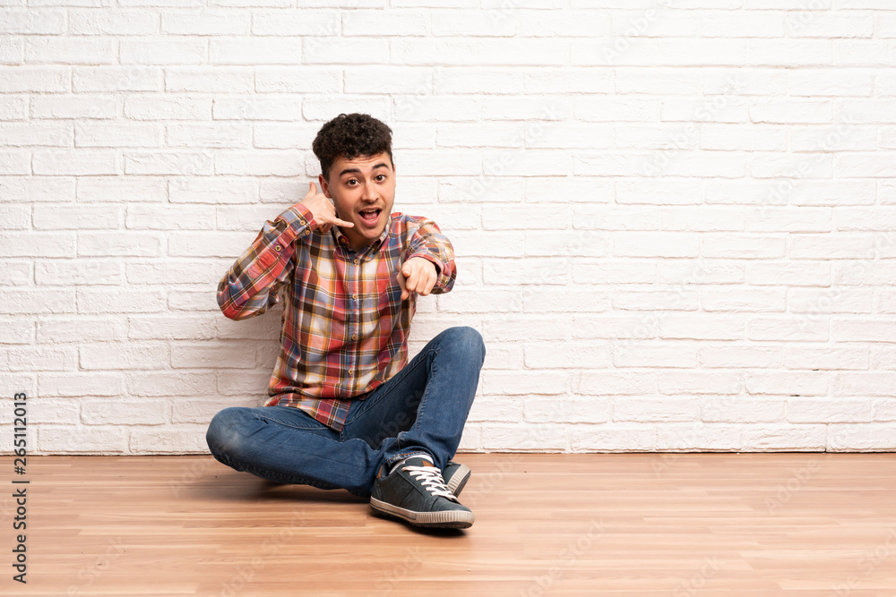 Young man sitting on the floor making phone gesture and pointing front