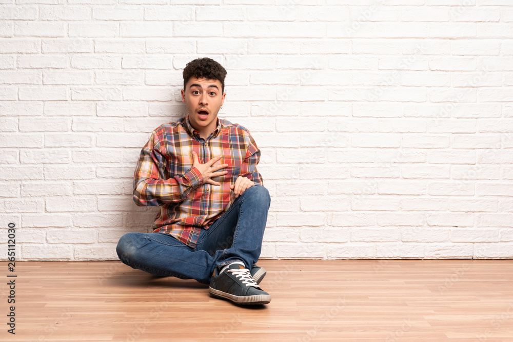 Young man sitting on the floor surprised and shocked while looking right Young man sitting on the floor surprised and shocked while looking right