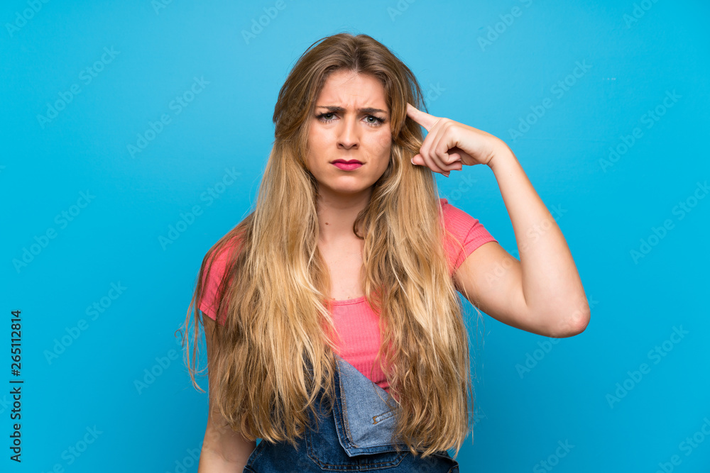Young blonde woman with overalls over isolated blue wall making the gesture of madness putting finger on the head