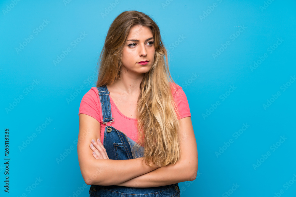 Young blonde woman with overalls over isolated blue wall portrait Young blonde woman with overalls over isolated blue wall portrait