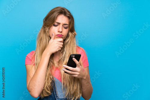 Young blonde woman with overalls over isolated blue wall thinking and sending a message