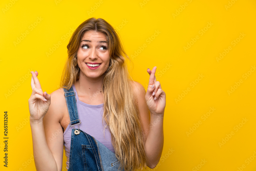 Young blonde woman with overalls over isolated yellow background with fingers crossing Young blonde woman with overalls over isolated yellow background with fingers crossing