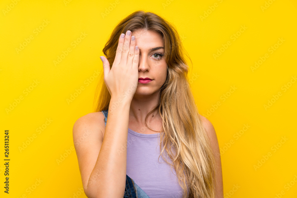 Young blonde woman with overalls over isolated yellow background covering a eye by hand Young blonde woman with overalls over isolated yellow background covering a eye by hand