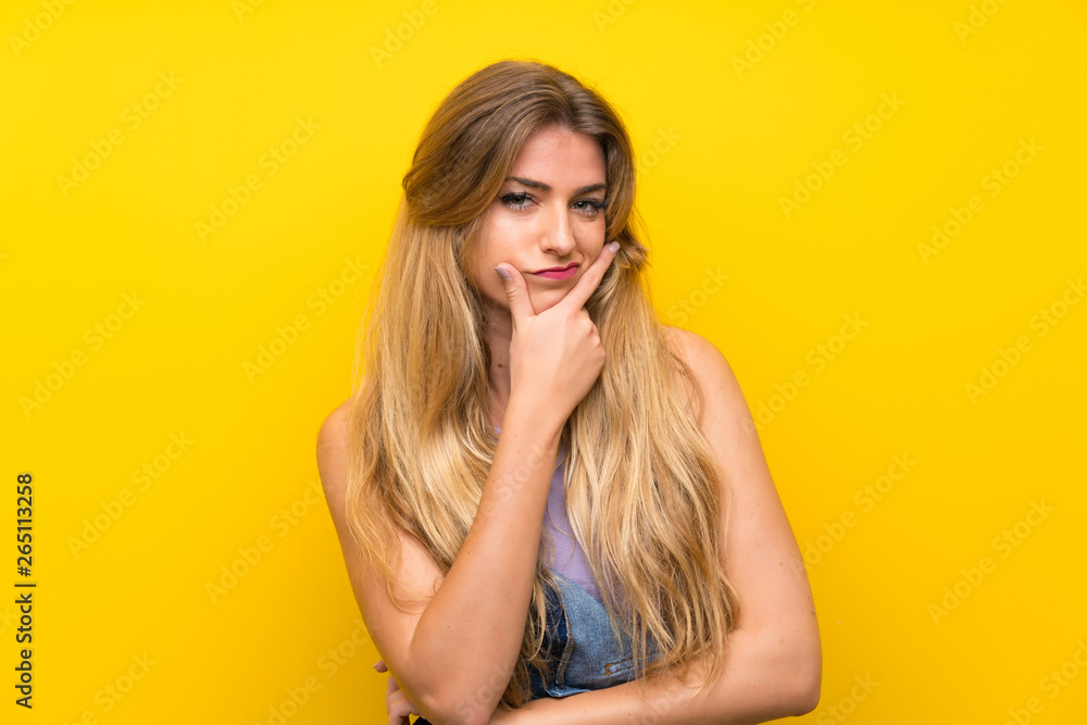 Young blonde woman with overalls over isolated yellow background standing and thinking an idea