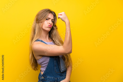 Young blonde woman with overalls over isolated yellow background making strong gesture