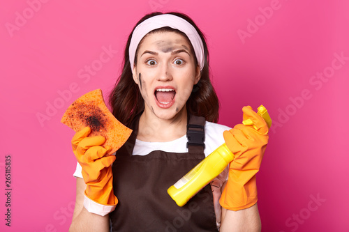 Impressed tired model posing isolated over pink background in studio, opening mouth with shock, doing household chores, wearing brown apron, t shirt and white headband. Housewife and cleaning concept. photo