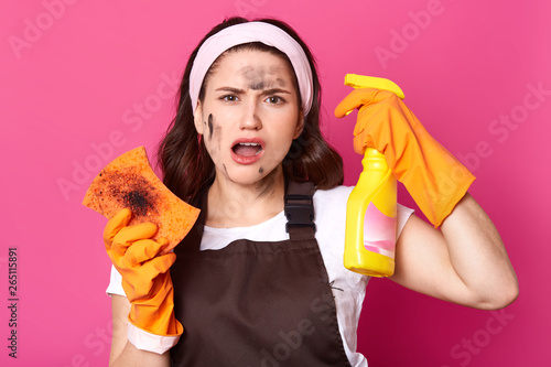 Hard working tired female opens her mouth widely with dissatisfaction, having dirty face and orange sponge, holding yellow bottle with cleaning liquid, wearing protective orange gloves for washing. photo