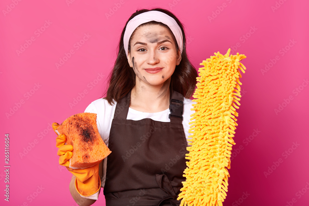 Slender adorable attractive girl looks directly at camera with bright broom and sponge in both hands, having dirt on her face, wearing dark brown apron, t shirt and headband. Household chores concept. Slender adorable attractive girl looks directly at camera with bright broom and sponge in both hands, having dirt on her face, wearing dark brown apron, t shirt and headband. Household chores concept.