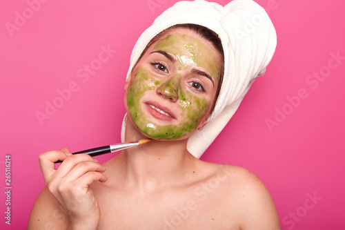 Beautiful delighted female looks directly at camera holding prof brush in one hand, putting light green mask with its help, waiting for good result, having white towel on head, standing naked.