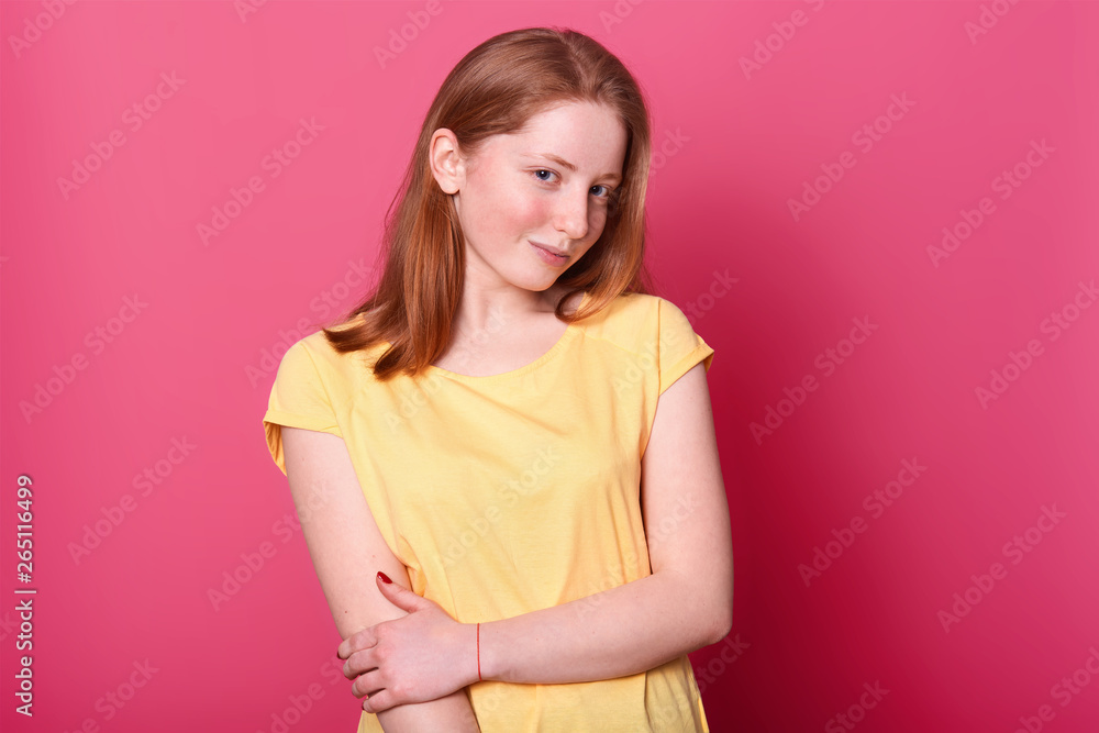 Charming magnetic positive girl poses isolated over bright pink background in studio, holding her arm, looking mysterious at camera. Talented sweet model wearing casual light yellow t shirt. Charming magnetic positive girl poses isolated over bright pink background in studio, holding her arm, looking mysterious at camera. Talented sweet model wearing casual light yellow t shirt.