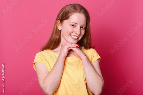 Attractive young sweet cute girl with smile on her face, holding arms near chin. Red haired lady with red manicure poses in studio isolated over pink background. People and emotions concept.