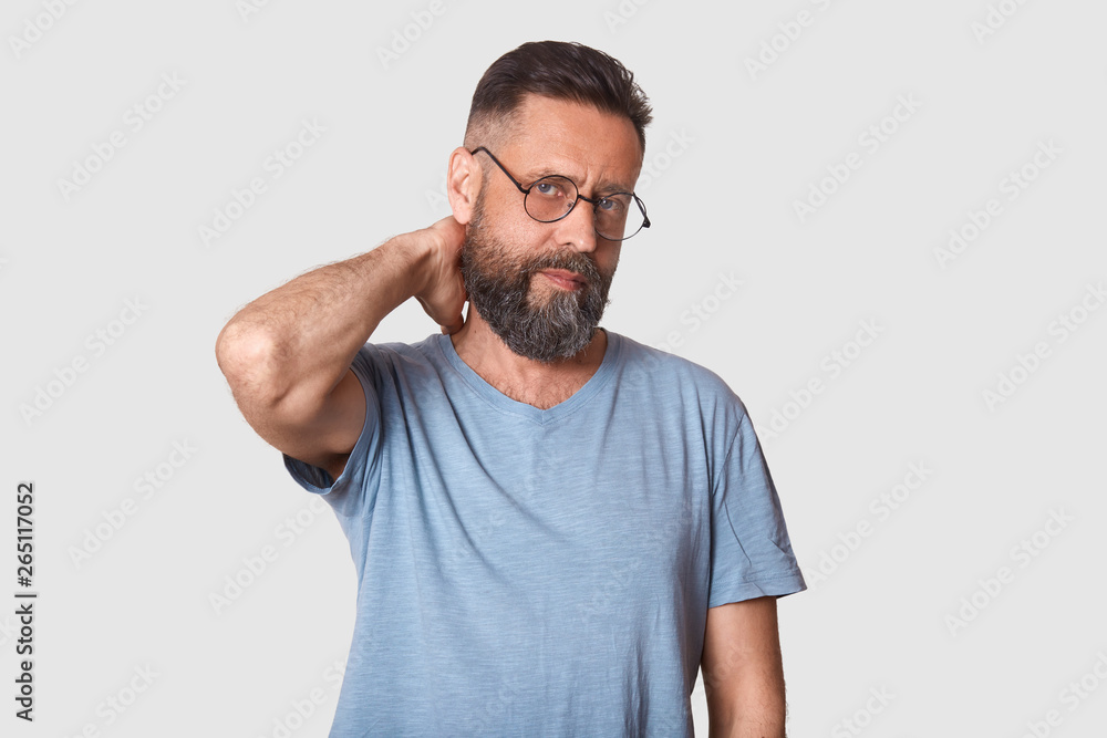 Tired unemotional handsome executive looks firmly at camera, holding his arm behind, making neck massage after hard working day. Middle aged black haired model wearing stylish specs and t shirt. Tired unemotional handsome executive looks firmly at camera, holding his arm behind, making neck massage after hard working day. Middle aged black haired model wearing stylish specs and t shirt.