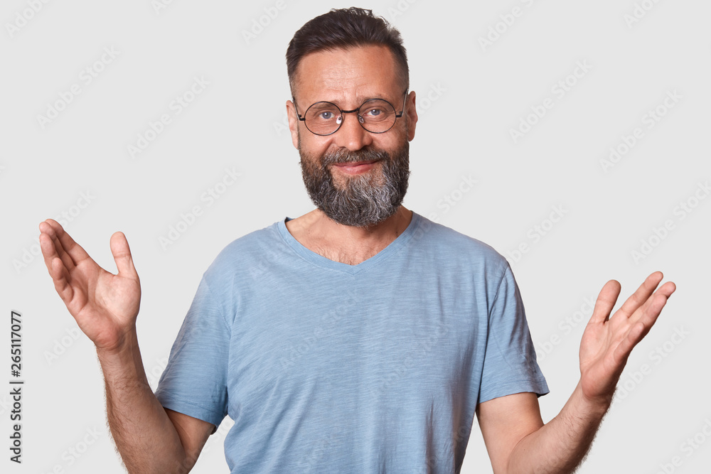 Smiling peaceful man rises his arms, greets somebody, looks pleased, wearing trendy spectacles and blue casual t shirt, spends spare time with pleasure. People, emotions and communication concept. Smiling peaceful man rises his arms, greets somebody, looks pleased, wearing trendy spectacles and blue casual t shirt, spends spare time with pleasure. People, emotions and communication concept.